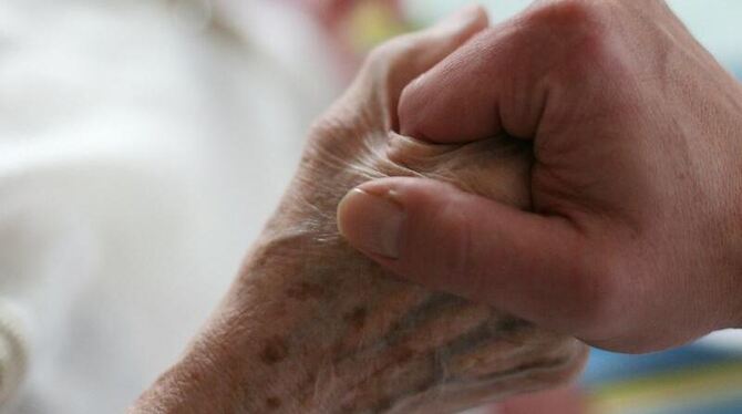 Ein Pfleger hält die Hand einer alten, kranken Frau, die als Pflegefall bettlägerig ist. Foto: Oliver Berg / Archiv