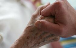 Ein Pfleger hält die Hand einer alten, kranken Frau, die als Pflegefall bettlägerig ist. Foto: Oliver Berg / Archiv