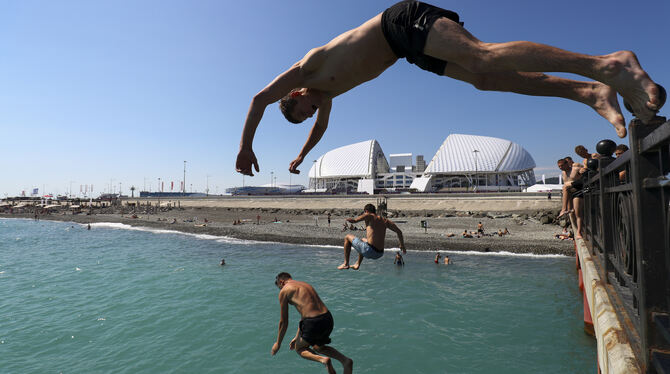 Junge Männer springen in Sotchi von einer Seebrücke an der Schwarzmeerküste von Adler. Im Hintergrund ist das Olympiastadion "Fi