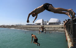 Junge Männer springen in Sotchi von einer Seebrücke an der Schwarzmeerküste von Adler. Im Hintergrund ist das Olympiastadion "Fi