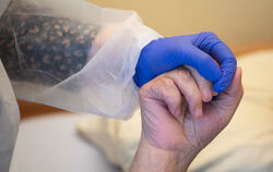 Eine Altenpflegerin in Schutzausrüstung hält die Hand eines Bewohners. FOTO: DPA