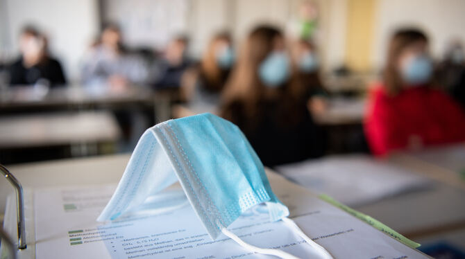 ine Maske liegt im Unterricht auf Unterlagen, während im Hintergrund Schülerinnen und Schüler in einem Gymnasium mit Mund- und N