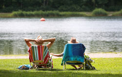 Ein Mann und eine Frau mit Strohhüten sitzen an einem Badesee in ihren Stühlen in der Sonne.
