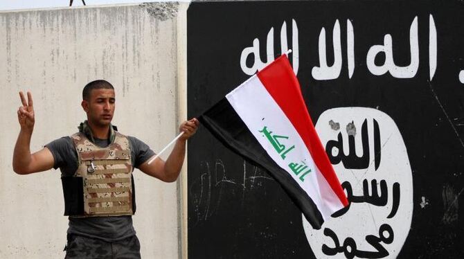 Ein irakischer Soldat in Tikrit steht mit seiner Landesflagge vor einer Wand mit dem Emblem der Terrororganisation IS. Foto: